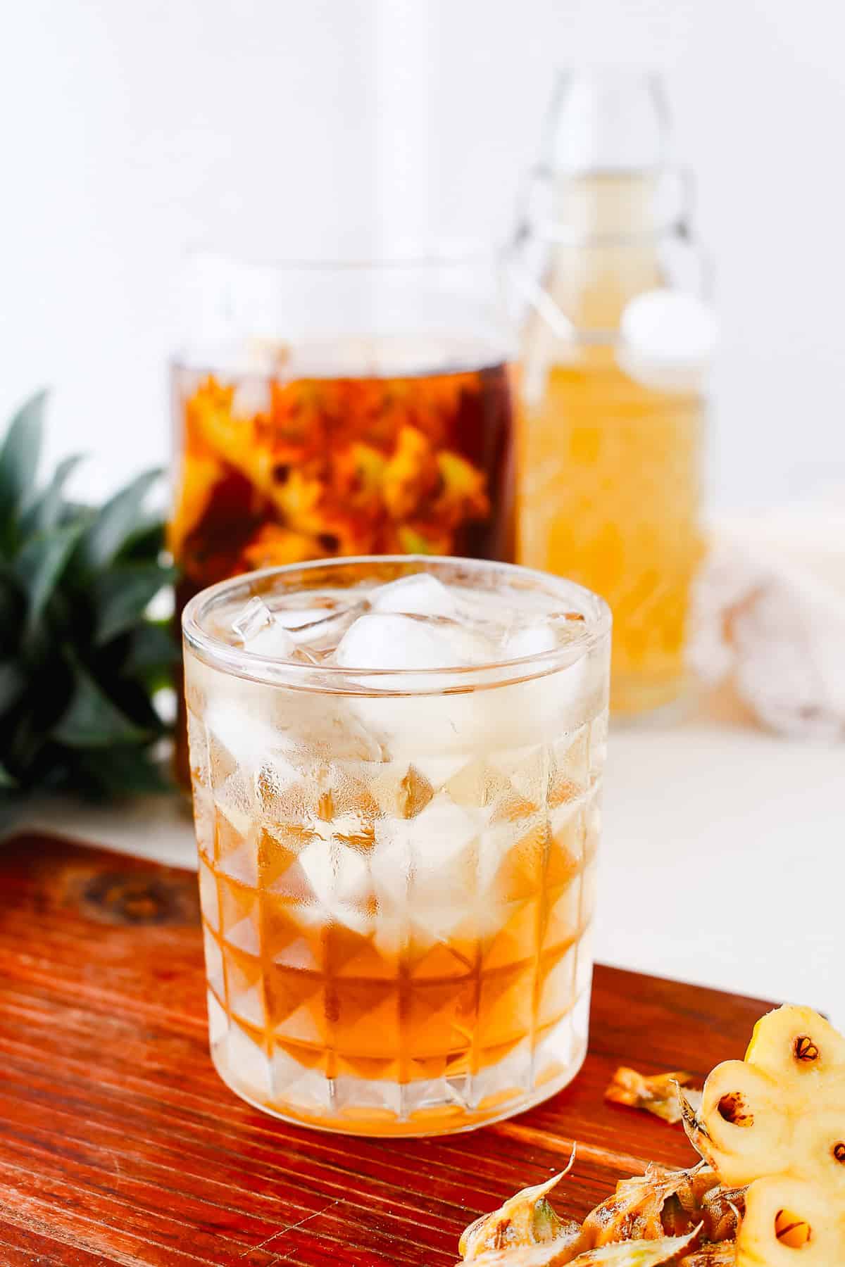 A glass of iced pineapple cocktail on a wooden surface, made with a vibrant fermented pineapple base, sits beside fresh pineapple and bottles in the background.