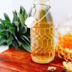 A glass bottle of pineapple vinegar on a wooden board, with pineapple leaves and peels beside itโa perfect ingredient for your next tepache recipe or other fermented drink creations.