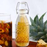 A glass bottle of light yellow fermented drink sits beside a jar with pineapple peels and a fresh pineapple on a wooden board, perfect for trying your own tepache recipe.