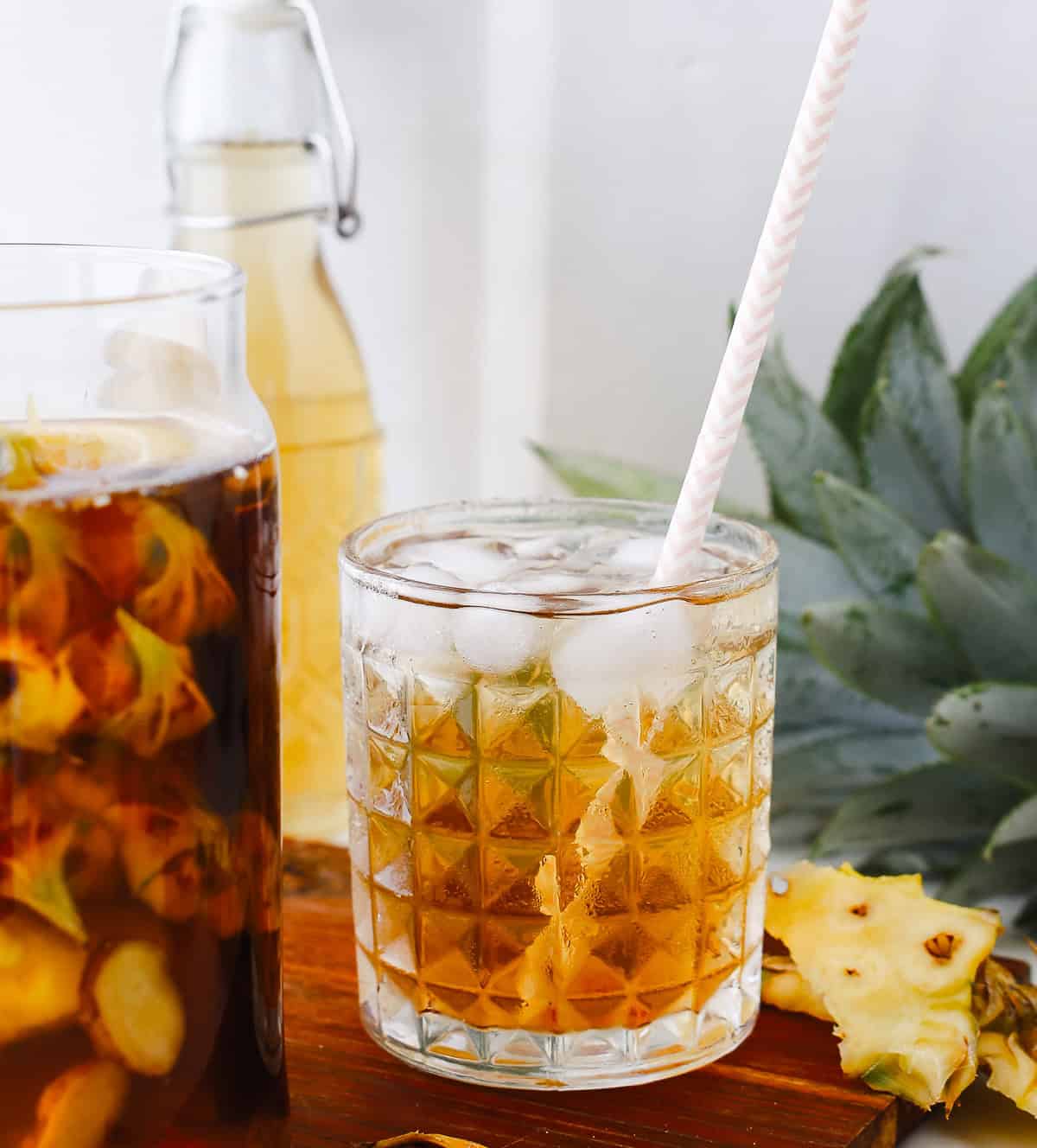 A glass of iced drink with a straw, pineapple slices, and a pineapple in the background—perfect for showcasing a refreshing pineapple tepache or trying your own fermented pineapple tepache recipe.