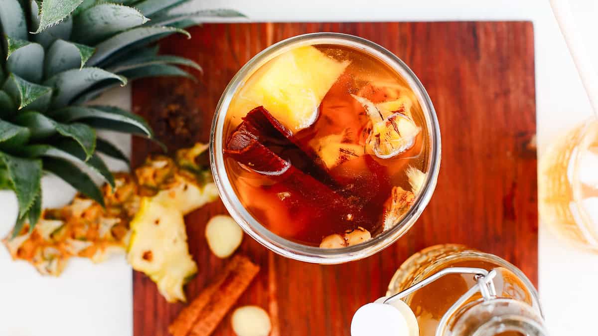 A glass of fermented pineapple drink sits on a wooden board with fresh pineapple, cinnamon, and nuts nearby—a classic tepache recipe perfect for refreshing sips.