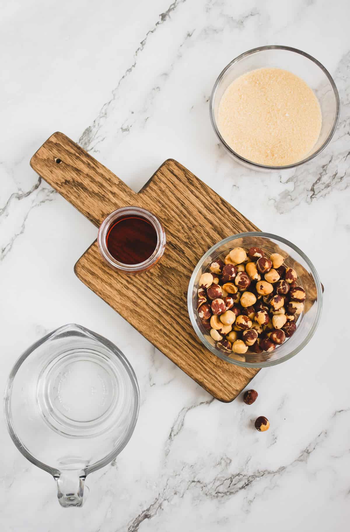 Bowls of hazelnuts, sugar, and liquid, plus a glass pitcher on a marble surface with a wooden board—perfect ingredients for crafting your own Homemade Hazelnut Syrup.