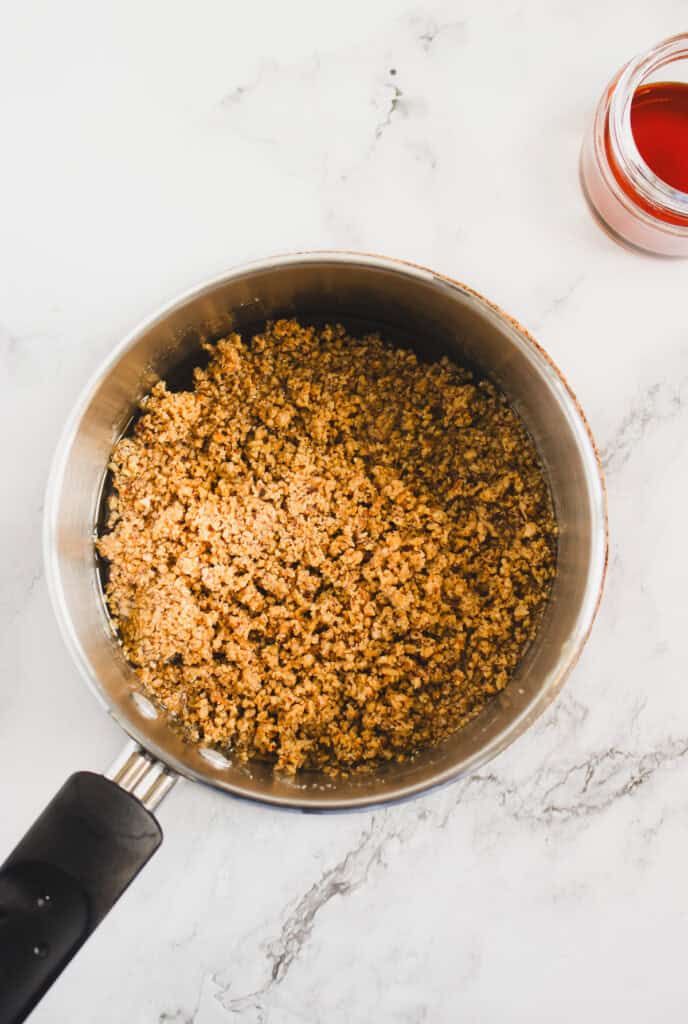 A saucepan filled with cooked ground meat sits on a marble surface beside a jar of homemade hazelnut syrup, its rich color hinting at the sweet, nutty flavor inside.