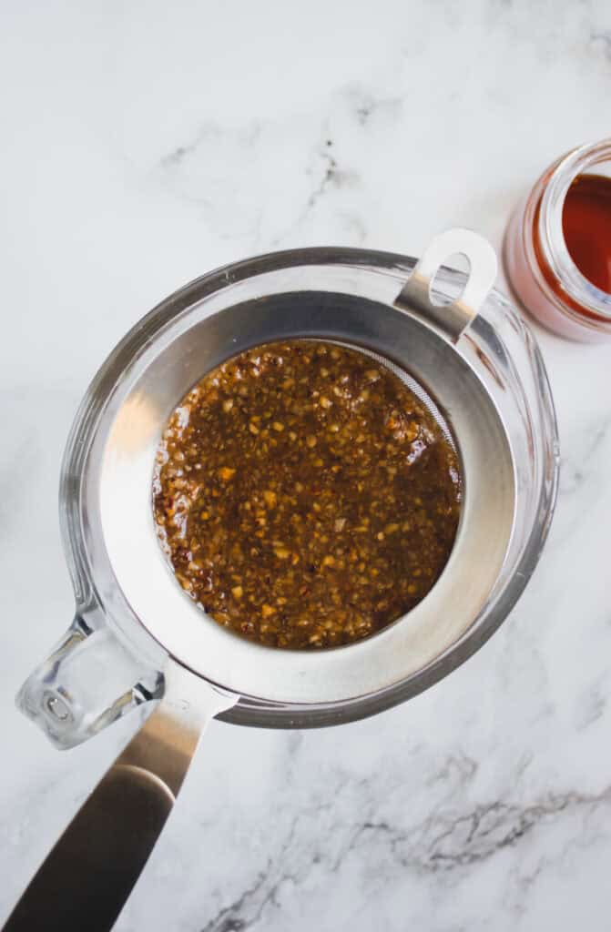 A metal strainer with a brown, chunky liquid—likely homemade hazelnut syrup—over a glass measuring cup, next to a jar on a marble surface.