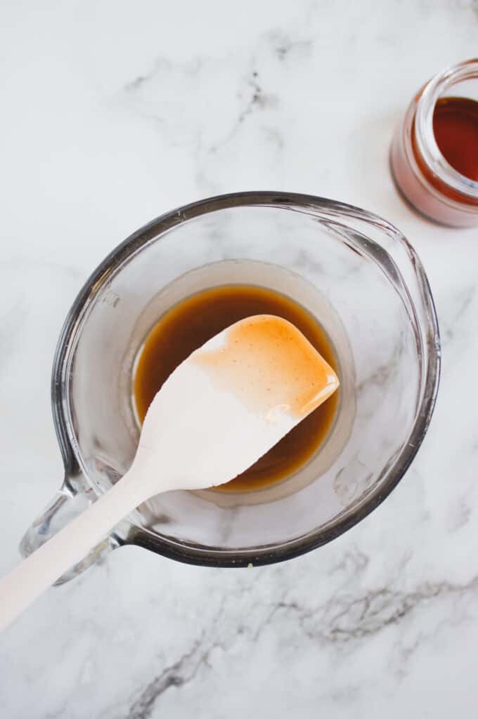 A white spatula rests in a glass measuring cup filled with homemade hazelnut syrup on a marble surface, ready for your next recipe.