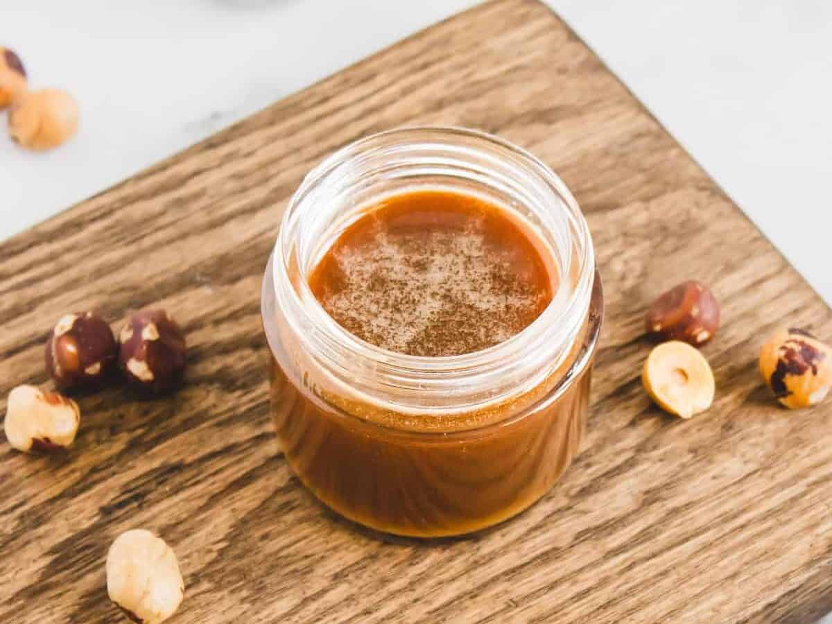 A jar of hazelnut spread on a wooden board, surrounded by whole and chopped hazelnuts, perfect for enjoying with your favorite homemade hazelnut syrup.