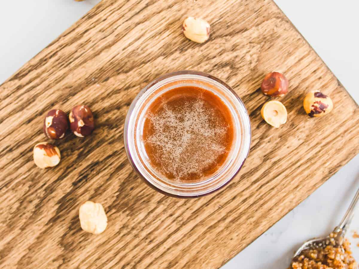 A jar of hazelnut spread on a wooden board, surrounded by whole hazelnuts and a spoonful of spread—perfect inspiration for your own Homemade Hazelnut Syrup or next Hazelnut Syrup Recipe.