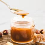 A spoon drizzles caramel-colored homemade hazelnut syrup into a small glass jar on a wooden surface, with hazelnuts nearby.