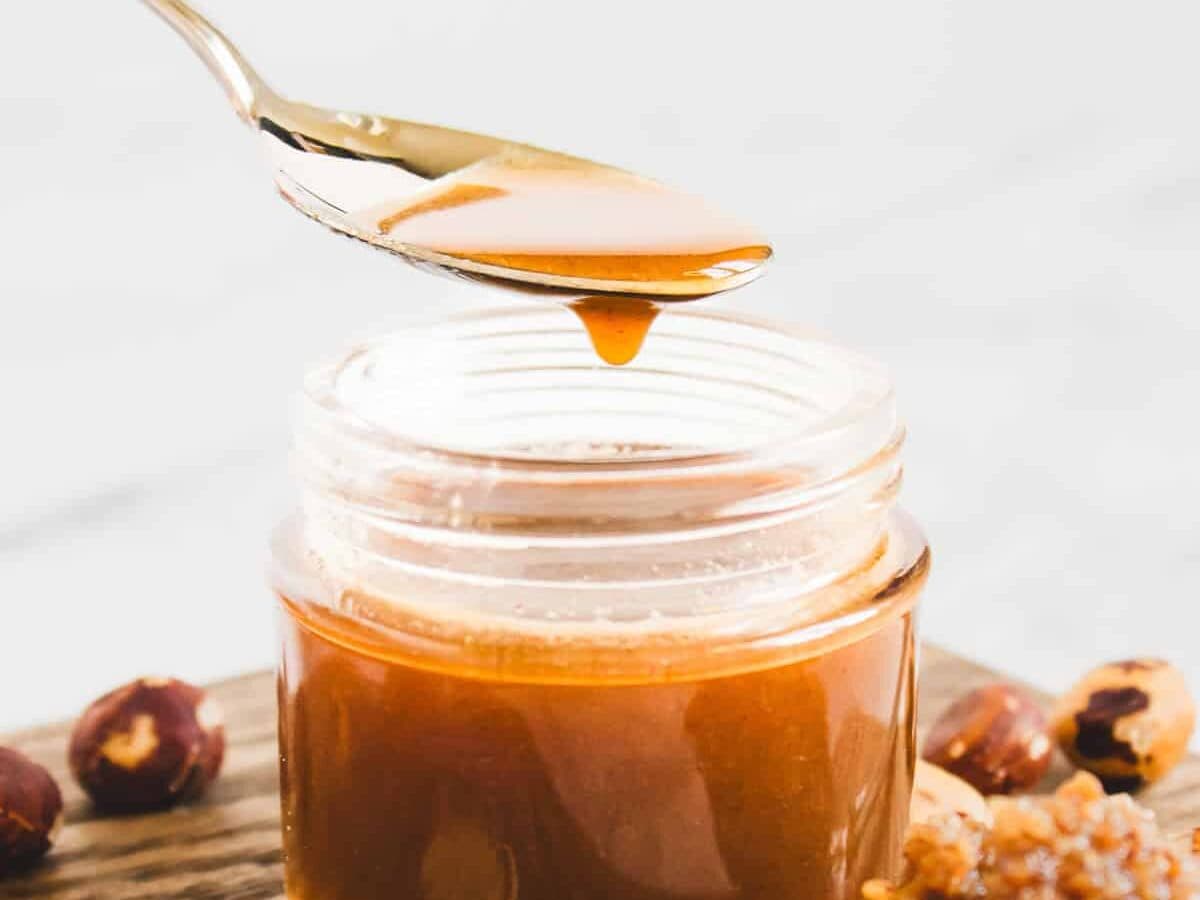 A spoon drizzles caramel-colored homemade hazelnut syrup into a small glass jar on a wooden surface, with hazelnuts nearby.