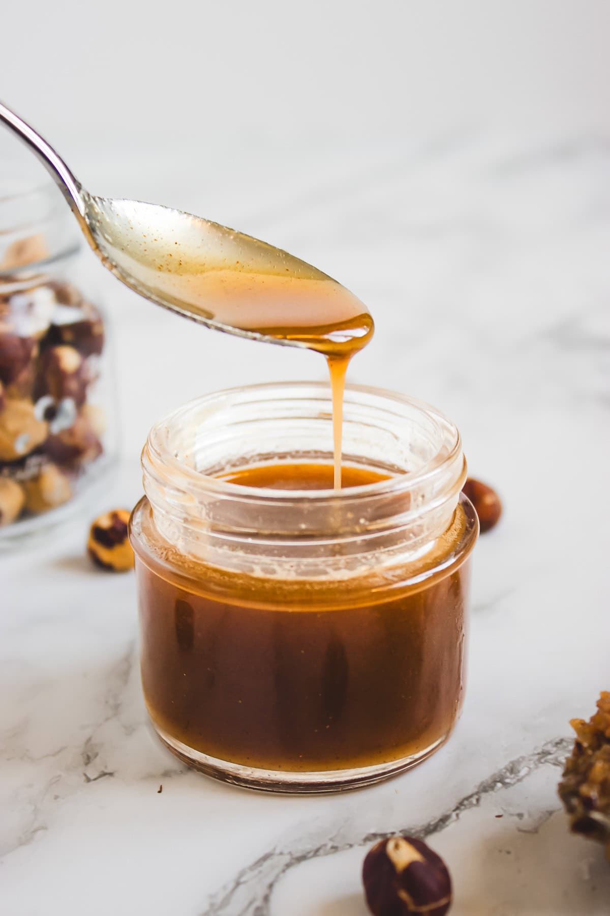 A spoon drizzling homemade caramel sauce into a small glass jar, with hazelnuts and a hint of hazelnut syrup on the marble surface nearby—perfect for your next dessert recipe.