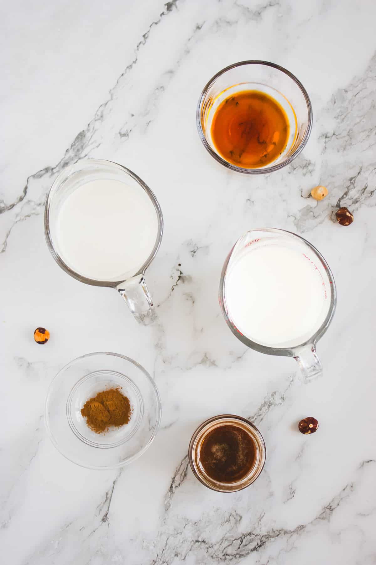 Five glass containers with milk, spices, and coffee creamer sit on a marble surface, surrounded by scattered hazelnuts.
