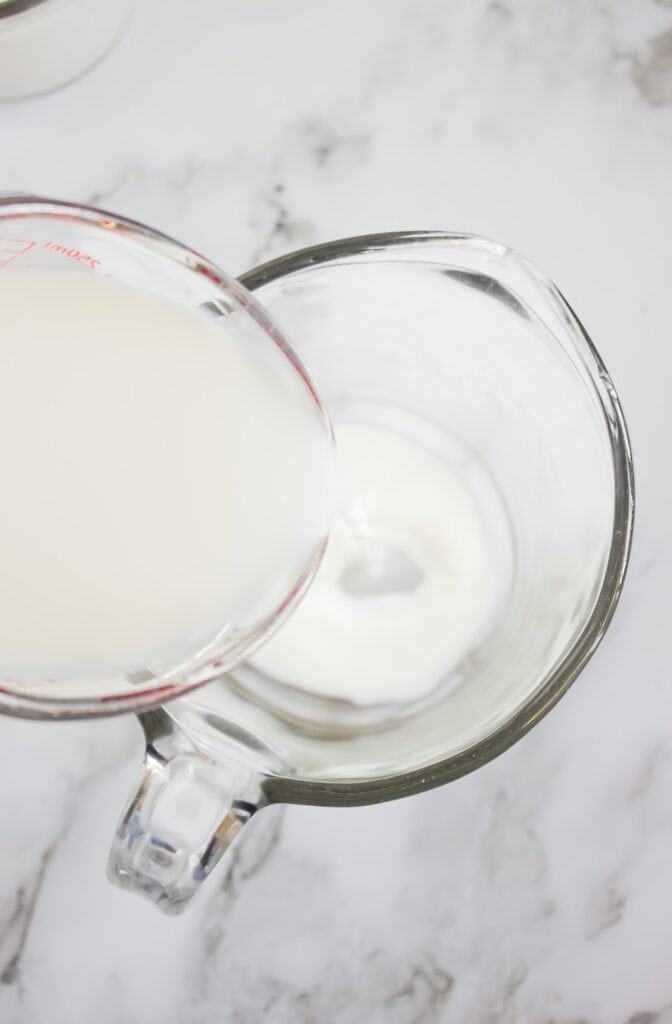 Milk and hazelnut coffee creamer being poured from a measuring cup into a glass pitcher on a white marble surface.