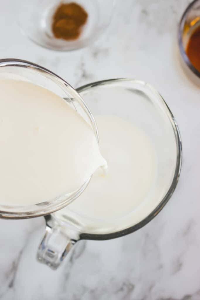 Creamy coffee creamer being poured from one glass measuring cup into another on a marble countertop.