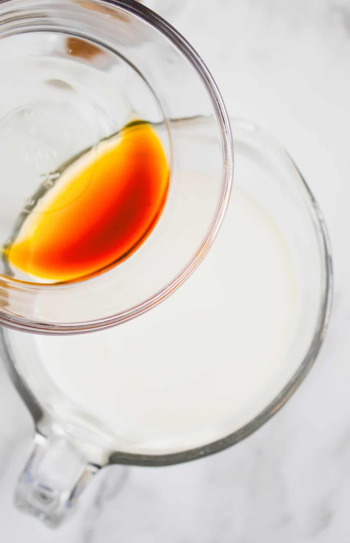 A glass bowl with hazelnut vanilla extract being poured into a larger bowl of milk on a white surface.
