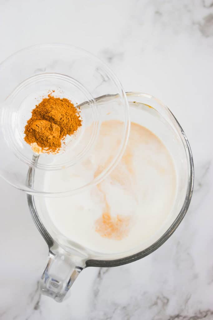 A glass bowl of ground spices held over a pitcher of milk on a marble surface, ready to transform it into a luscious Hazelnut Coffee Creamer.