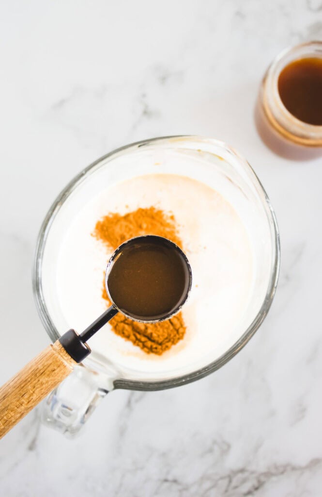 A measuring spoon with hazelnut coffee creamer held above a bowl of cream and cinnamon on a marble surface.