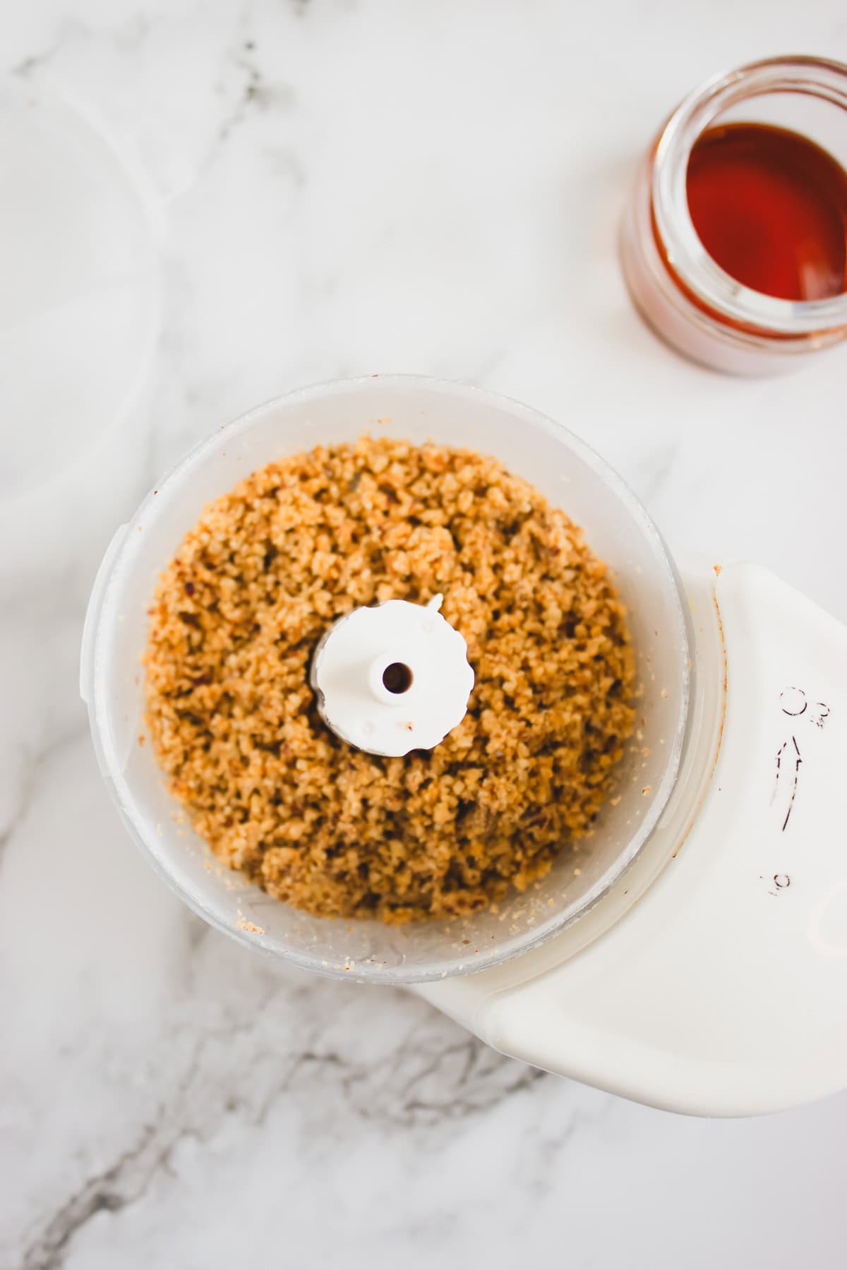 A food processor with chopped nuts inside sits next to a small jar of honey on a white marble surface, perfect for crafting a homemade recipe or delicious hazelnut syrup.