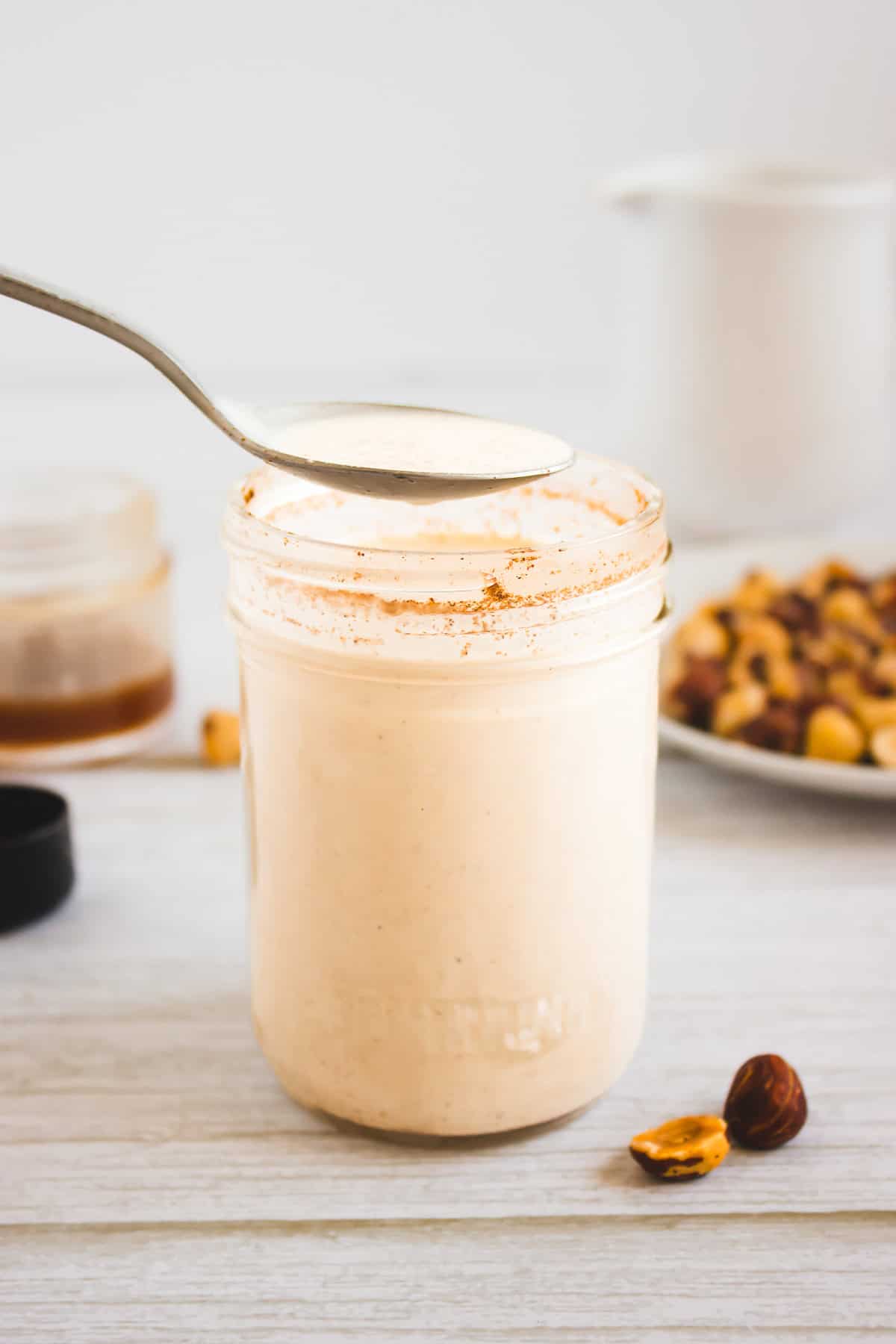 A spoon above a jar of creamy hazelnut smoothie, with nuts and honey in the background on a white surface.