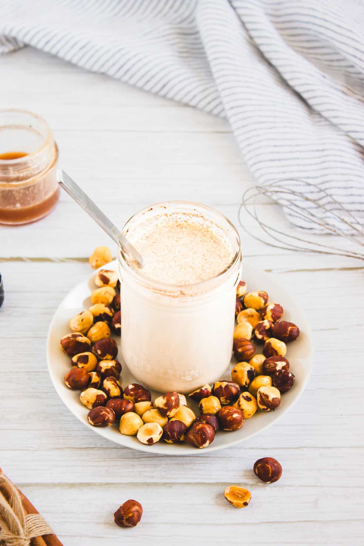 A glass jar of creamy hazelnut coffee creamer with a spoon, surrounded by hazelnuts on a white plate, on a light table.