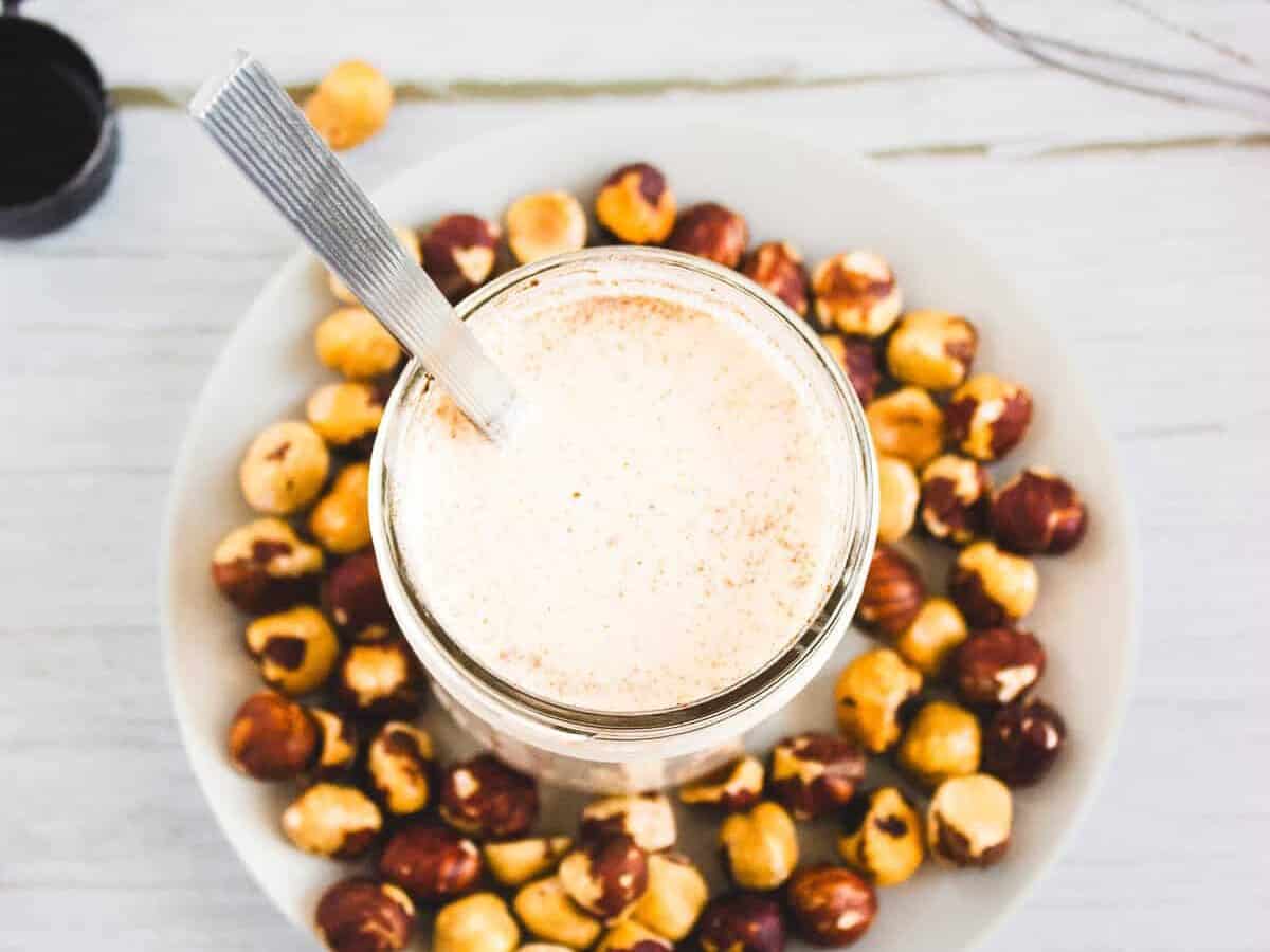 Glass of frothy hazelnut milk on a plate of hazelnuts, with a spoon and a jar of honey on a white table—perfect as a Hazelnut Coffee Creamer for your morning brew.