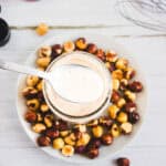 A spoonful of creamy coffee creamer sauce over a jar, with a plate of hazelnuts on a white wooden table.