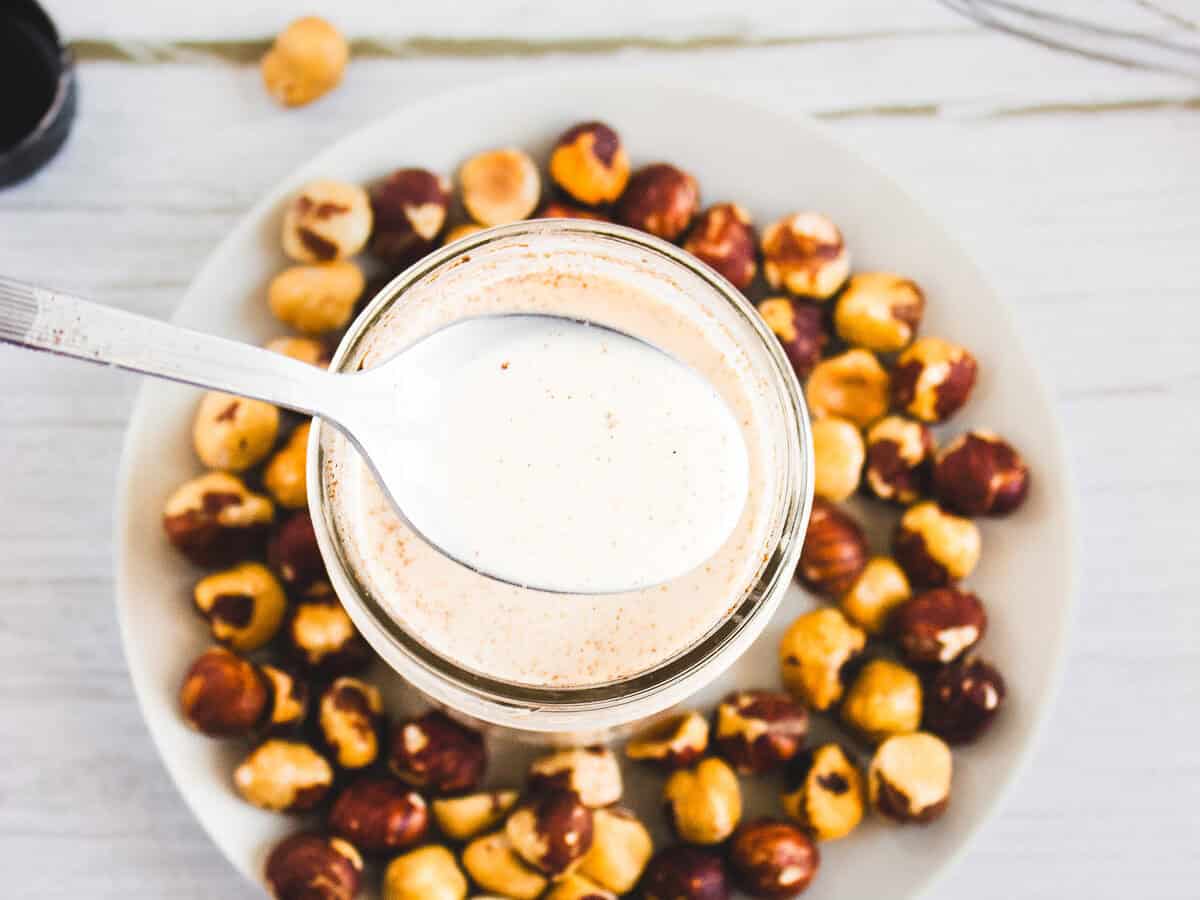 A spoonful of creamy coffee creamer sauce over a jar, with a plate of hazelnuts on a white wooden table.