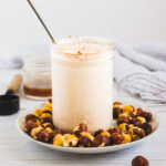 A glass jar of smoothie with a metal straw, surrounded by hazelnuts on a white plate-perfect for pairing with your favorite hazelnut coffee creamer.