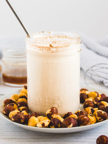 A glass jar of smoothie with a metal straw, surrounded by hazelnuts on a white plate—perfect for pairing with your favorite hazelnut coffee creamer.