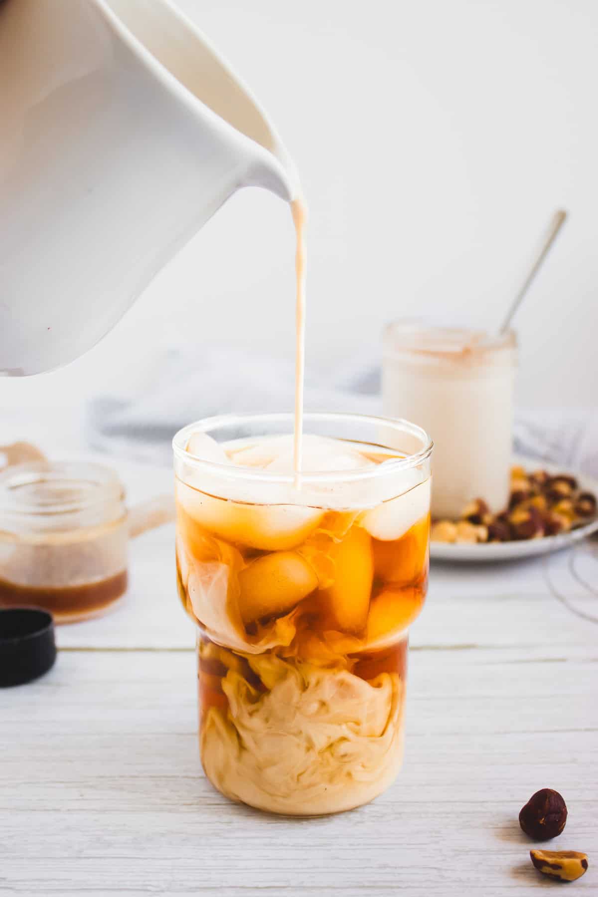 Creamy coffee creamer is poured into a glass of iced coffee, with jars and a plate of hazelnuts in the background, adding a hint of hazelnut flavor to the scene.