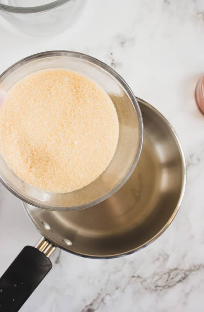 A glass bowl of granulated gelatin is held above a stainless steel saucepan on a marble countertop, ready to help craft a delicious Homemade Hazelnut Syrup.