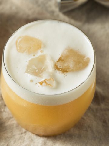 A round glass of frothy non-alcoholic tequila sour cocktail with ice cubes sits elegantly on a beige cloth surface.