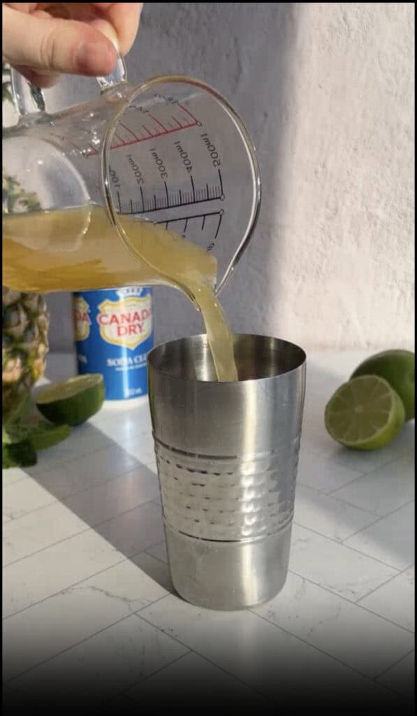 A hand pours juice from a measuring cup into a cocktail shaker for a refreshing pineapple mocktail recipe; limes and a soda can sit nearby.