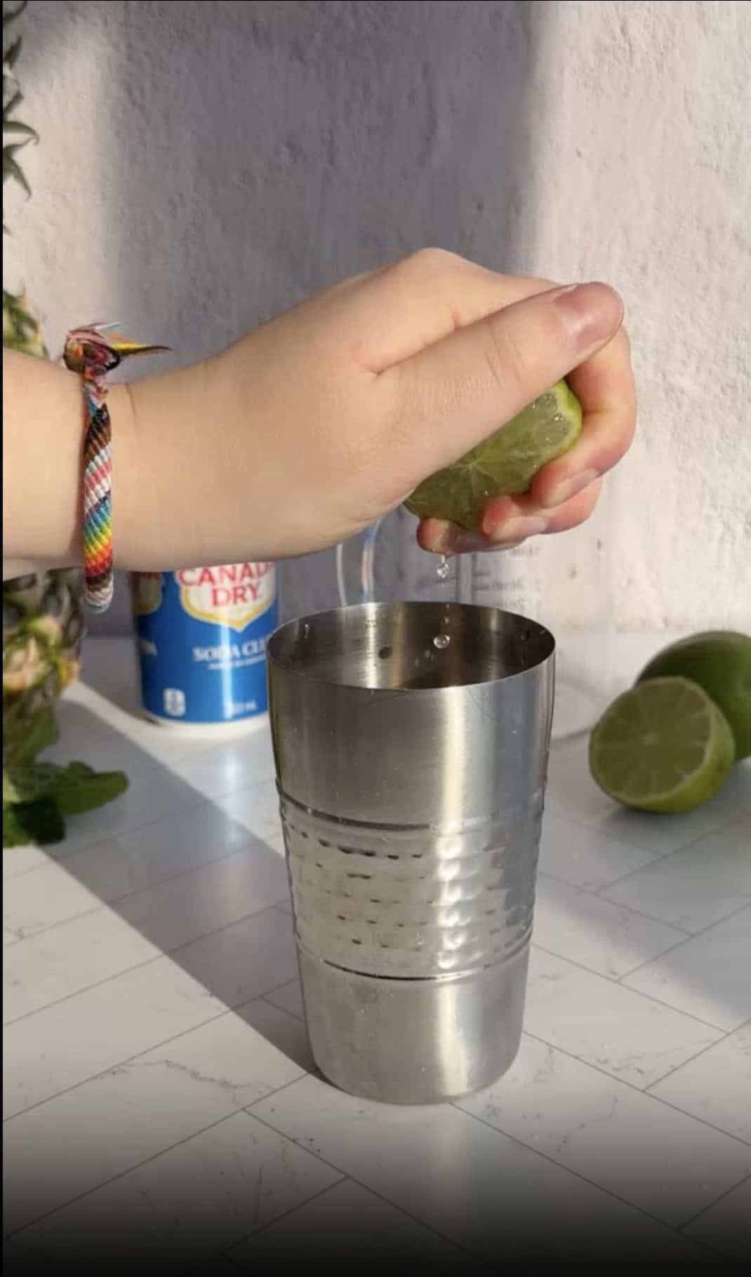 A hand squeezes a lime over a metal cocktail shaker on a white counter, preparing ingredients for a refreshing pineapple mocktail recipe.