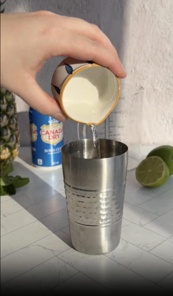 A hand pours liquid from a small cup into a metal shaker, with lime, pineapple, and soda in the background—perfect for crafting a refreshing pineapple mocktail recipe.