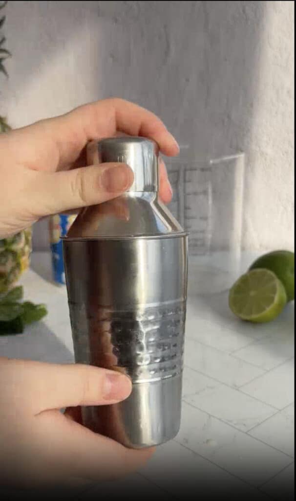 Hands holding a stainless steel cocktail shaker on a kitchen counter with limes, a measuring cup, and ingredients for a refreshing pineapple mocktail recipe nearby.