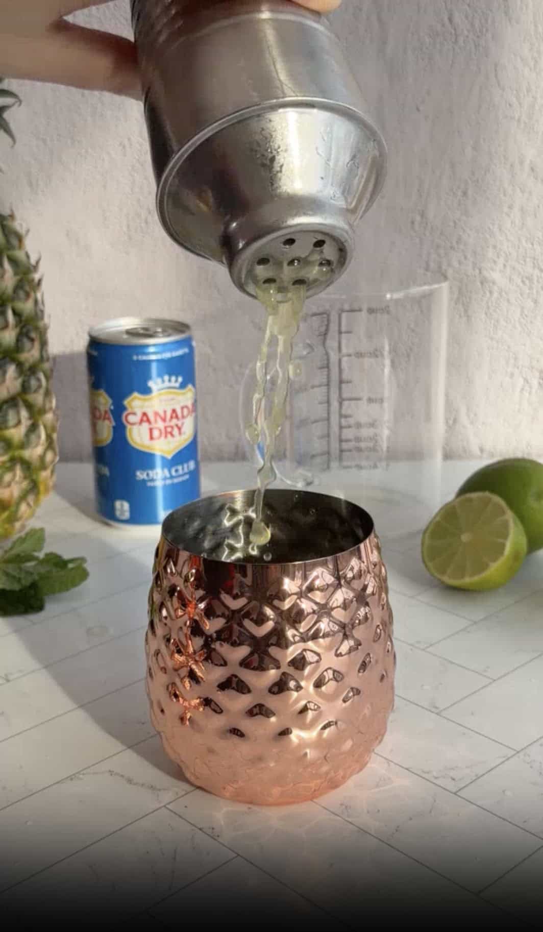 A cocktail shaker pours liquid into a copper cup, with a lime, soda can, and pineapple nearby—perfect for crafting your favorite pineapple mocktail recipe.