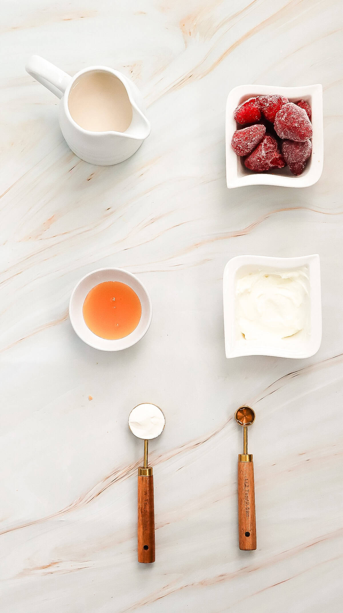 Flat lay of cream, frozen strawberries, honey, yogurt, and two wooden-handled scoops on a marbled surface&mdash;perfect ingredients for a Strawberry Cheesecake Frappuccino recipe inspired by the Starbucks copycat favorite.