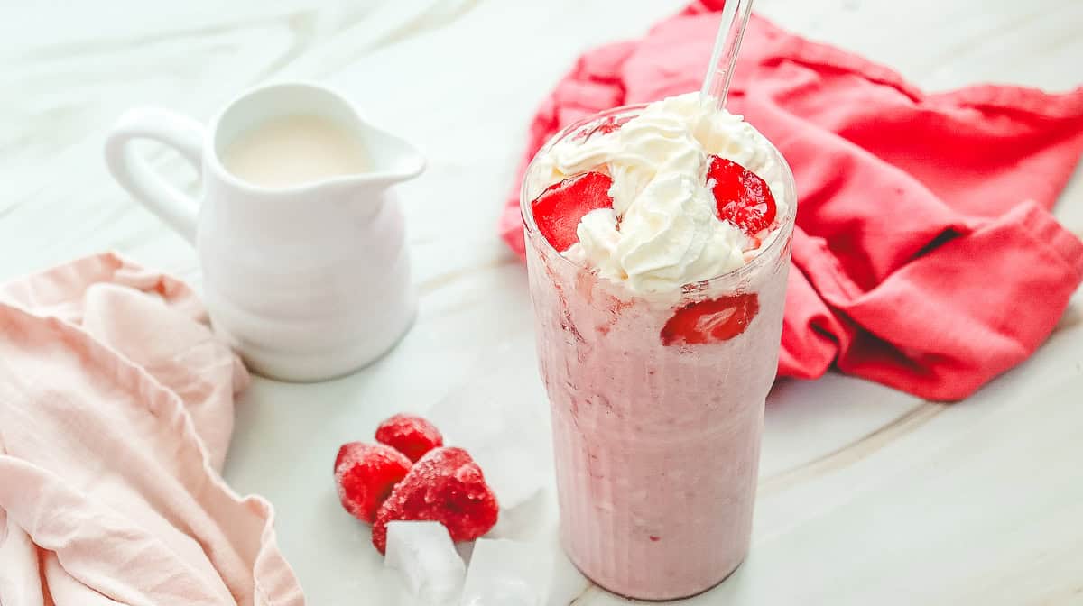A Starbucks Copycat strawberry milkshake topped with whipped cream, next to a small jug of milk and frozen strawberries&mdash;just like a homemade Strawberry Cheesecake Frappuccino recipe.