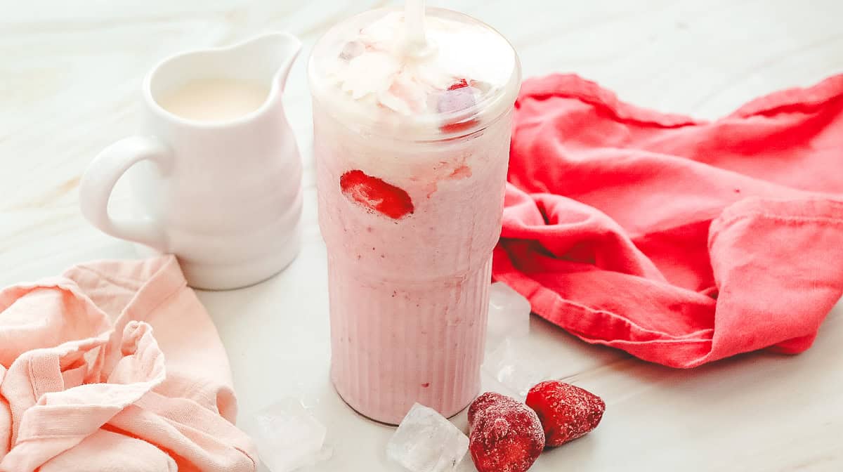A strawberry milkshake in a tall glass with frozen strawberries, ice cubes, and a small pitcher of milk nearby&mdash;perfect as a Starbucks Strawberry Cheesecake Frappuccino copycat recipe.
