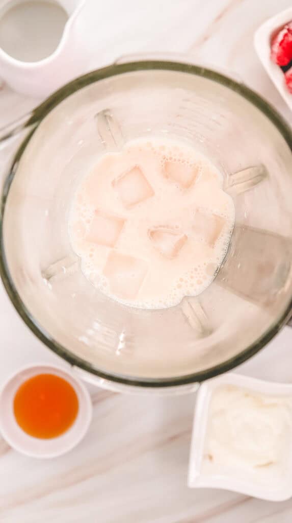 Top view of a blender with milk and ice cubes, surrounded by small bowls of ingredients on a white surface&mdash;perfect for crafting a Strawberry Cheesecake Frappucino Starbucks Copycat recipe.