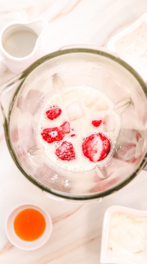 Top view of a blender with milk, strawberries, and ice, surrounded by small bowls of ingredients&mdash;perfect for making a Copycat Recipe of the Starbucks Strawberry Cheesecake Frappucino.