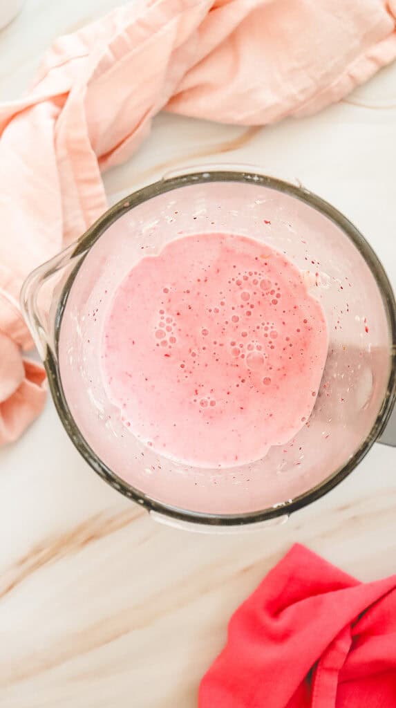 Top view of a blender with pink smoothie inside, resembling a Strawberry Cheesecake Frappuccino, surrounded by light and dark pink cloths on a marble surface&mdash;perfect for a Starbucks Copycat recipe at home.