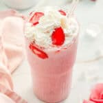 A pink strawberry milkshake inspired by a Starbucks Copycat Strawberry Cheesecake Frappuccino, topped with whipped cream and strawberries, served with a clear straw in a glass.