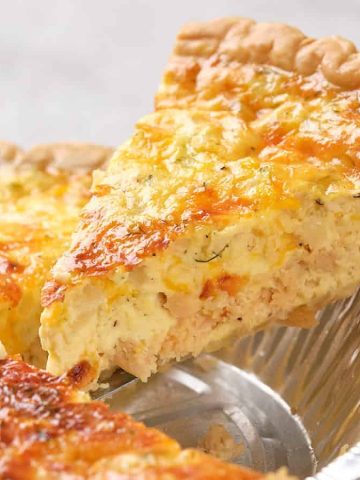 A hand lifts a slice of golden, cheesy quiche from a tin pie pan on a wooden board—perfect for hearty Father's Day breakfasts.