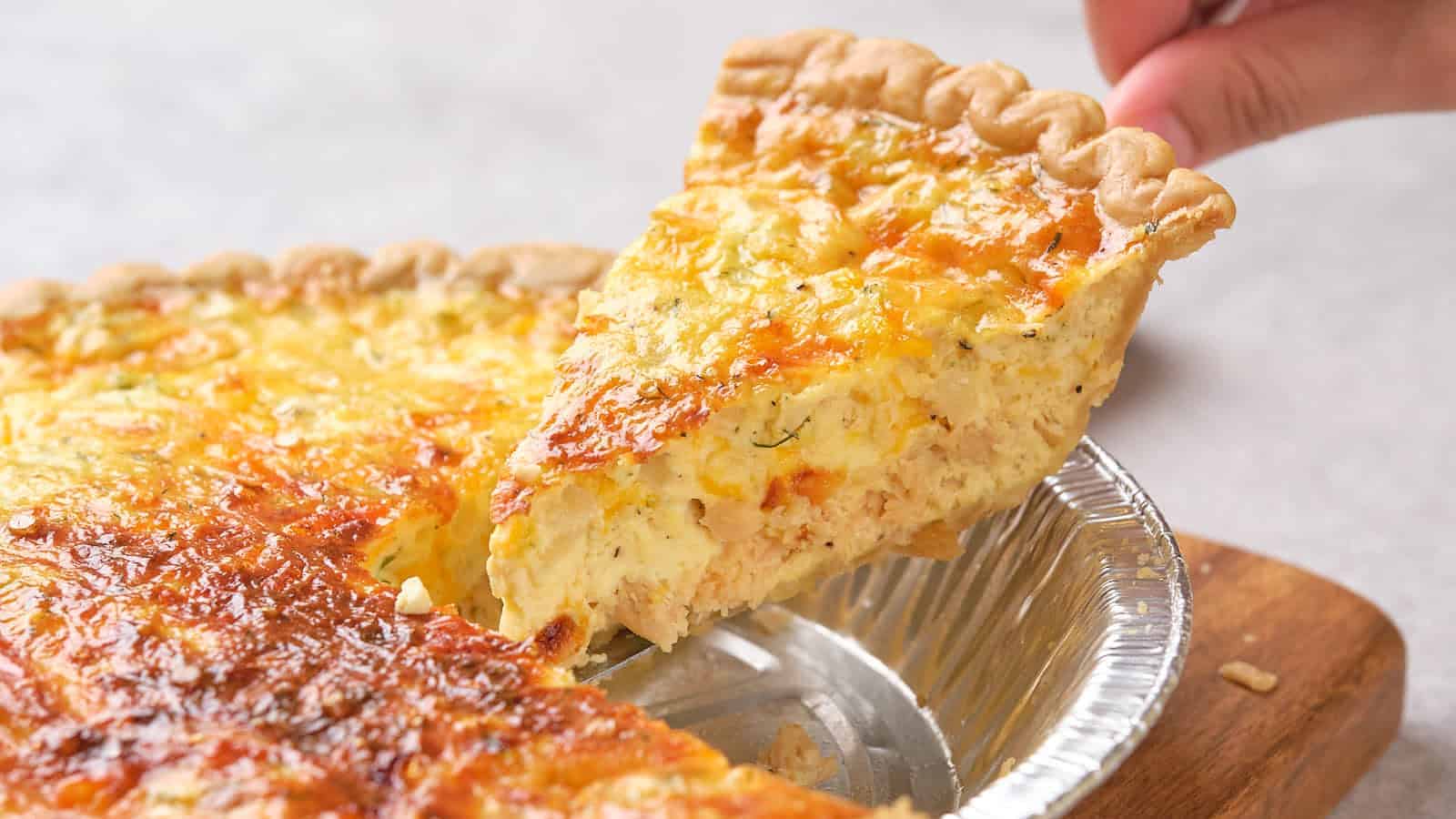 A hand lifts a slice of golden, cheesy quiche from a tin pie pan on a wooden board&mdash;perfect for hearty Father's Day breakfasts.