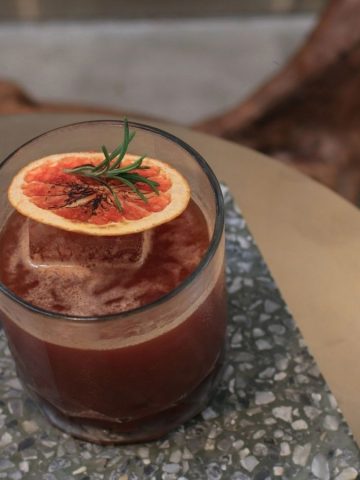 A dark mocktail garnished with a dried orange slice and rosemary, served on a terrazzo table—perfect for those seeking sophisticated non-alcoholic drinks.