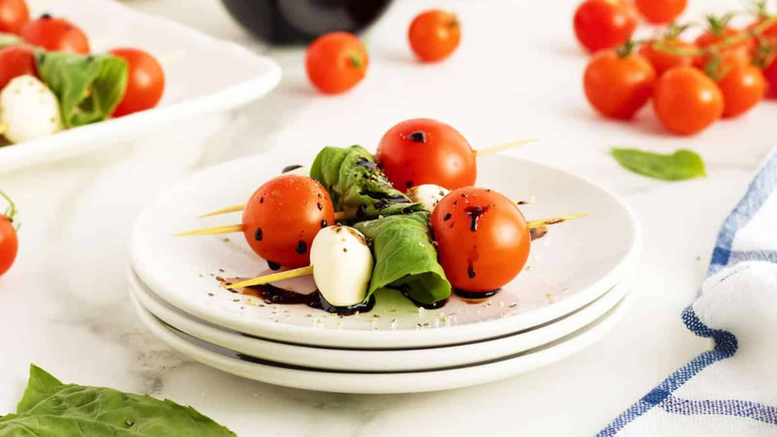Caprese skewers with tomatoes, mozzarella, basil, and balsamic glaze on a white plate with cherry tomatoes nearby.
