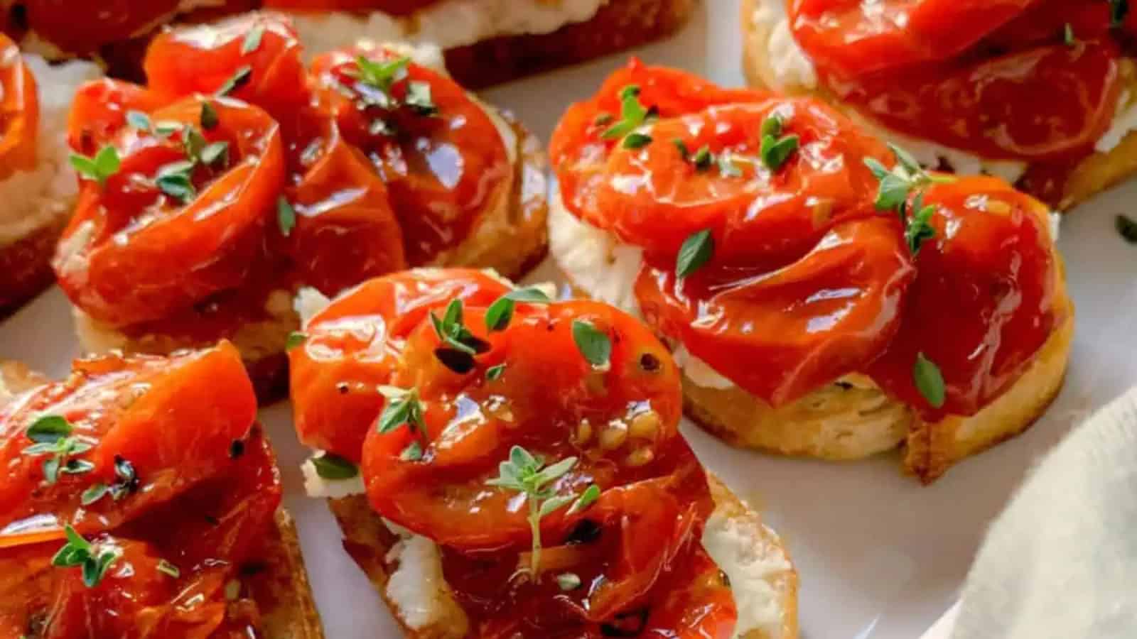 Slices of toasted bread topped with roasted tomatoes, creamy cheese, and fresh thyme on a white plate. The tomatoes are glistening, and the thyme adds a touch of green.