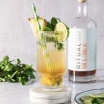 Tall glass with a pineapple mojito mocktail, lime, mint, and pineapple garnish; rum bottle in the background.