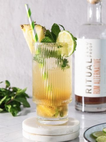 Tall glass with a pineapple mojito mocktail, lime, mint, and pineapple garnish; rum bottle in the background.